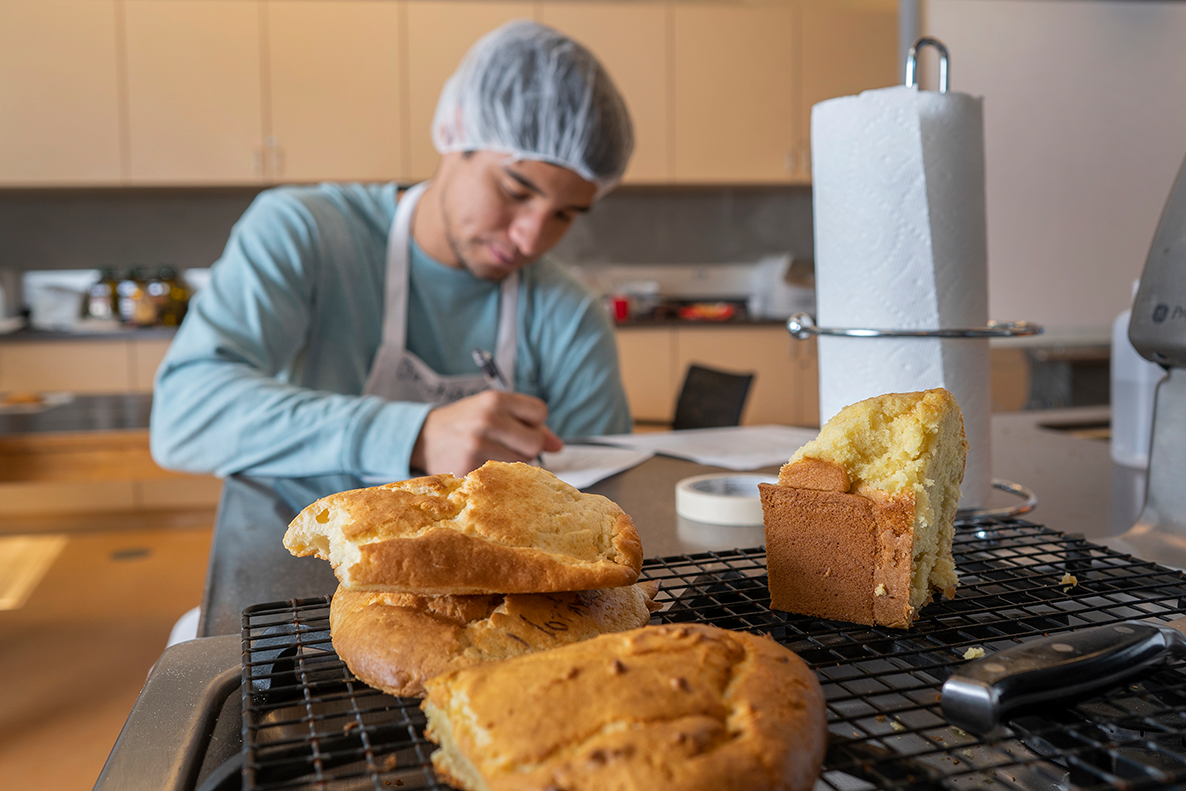 A person wearing an apron and hair net measures a cake.