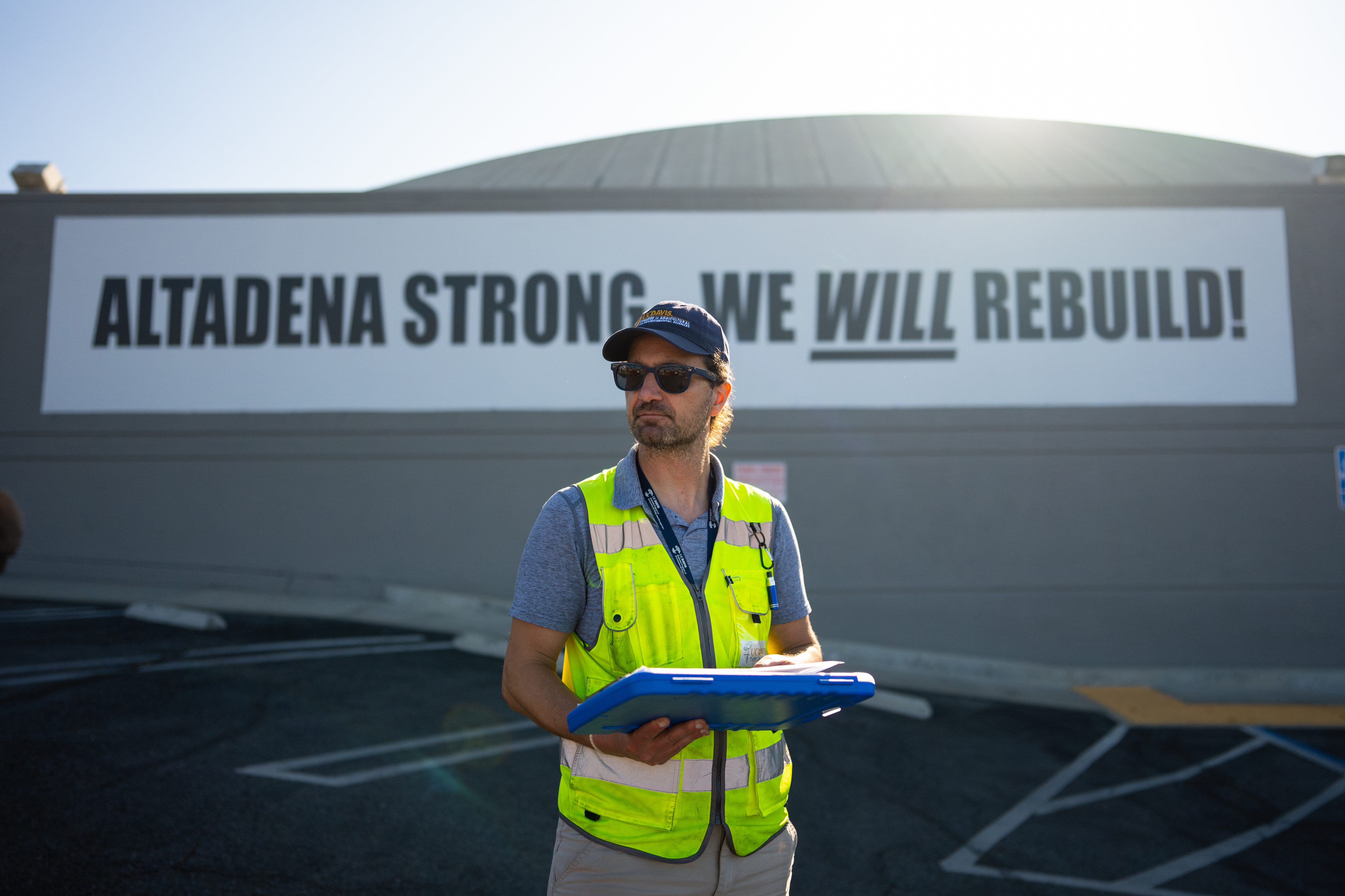 A man in neon vest and hard hat holding clipboard before Altadena Strong banner