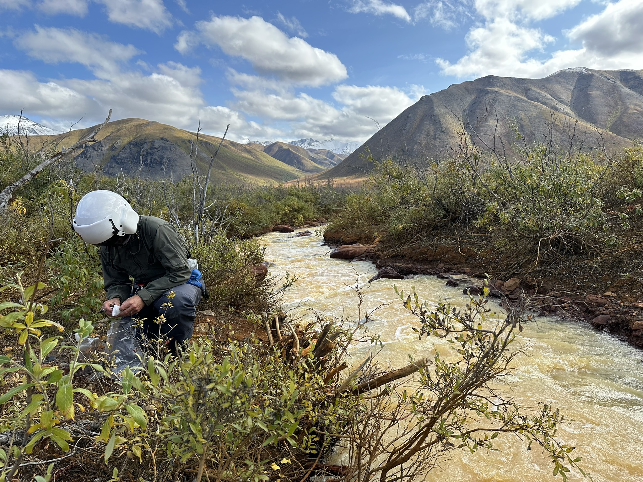 Person crouching by muddy stream in an alpine valley under a cloudy blue sky
