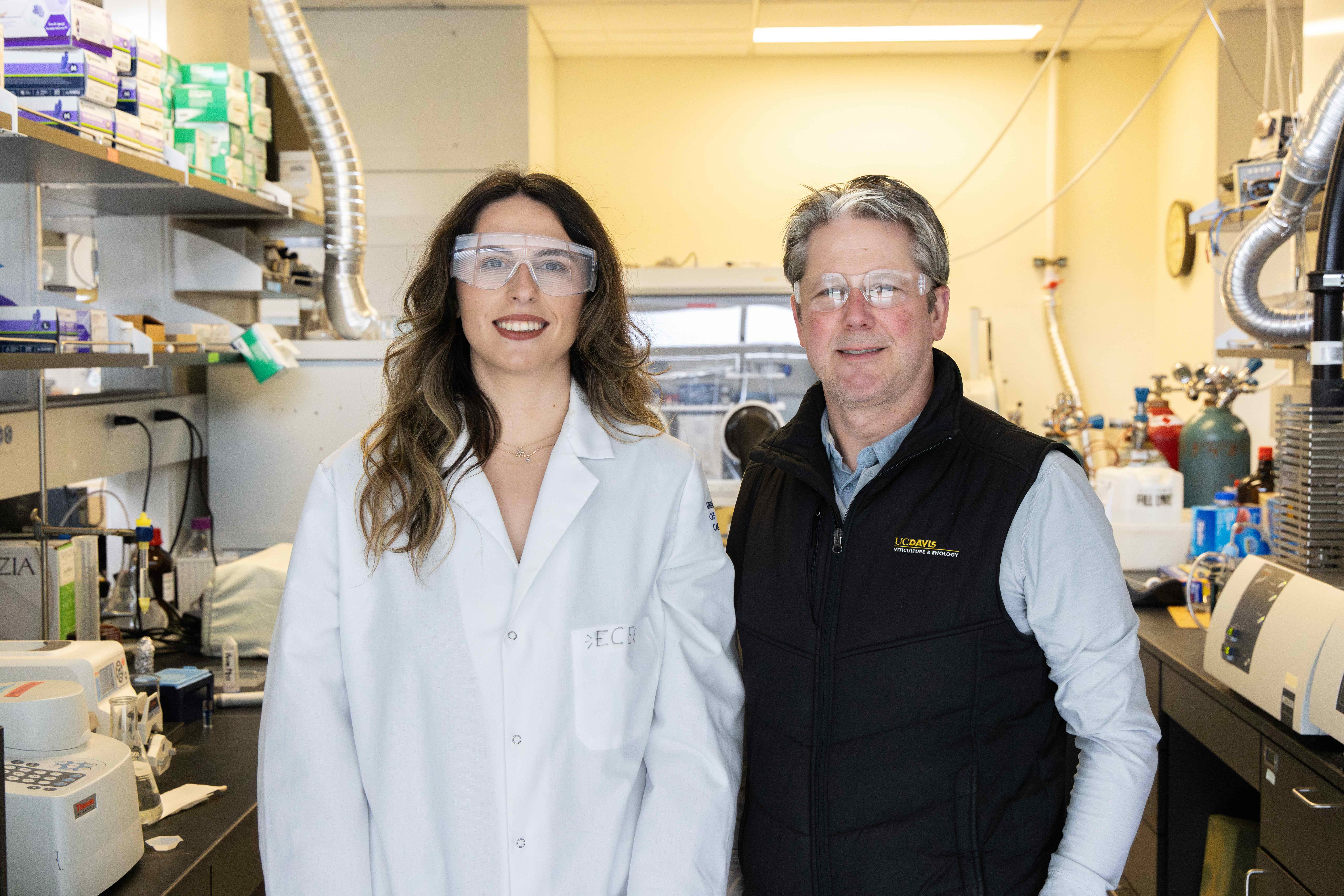 Smiling woman in lab coat and man in vest wearing safety glasses, laboratory photograph