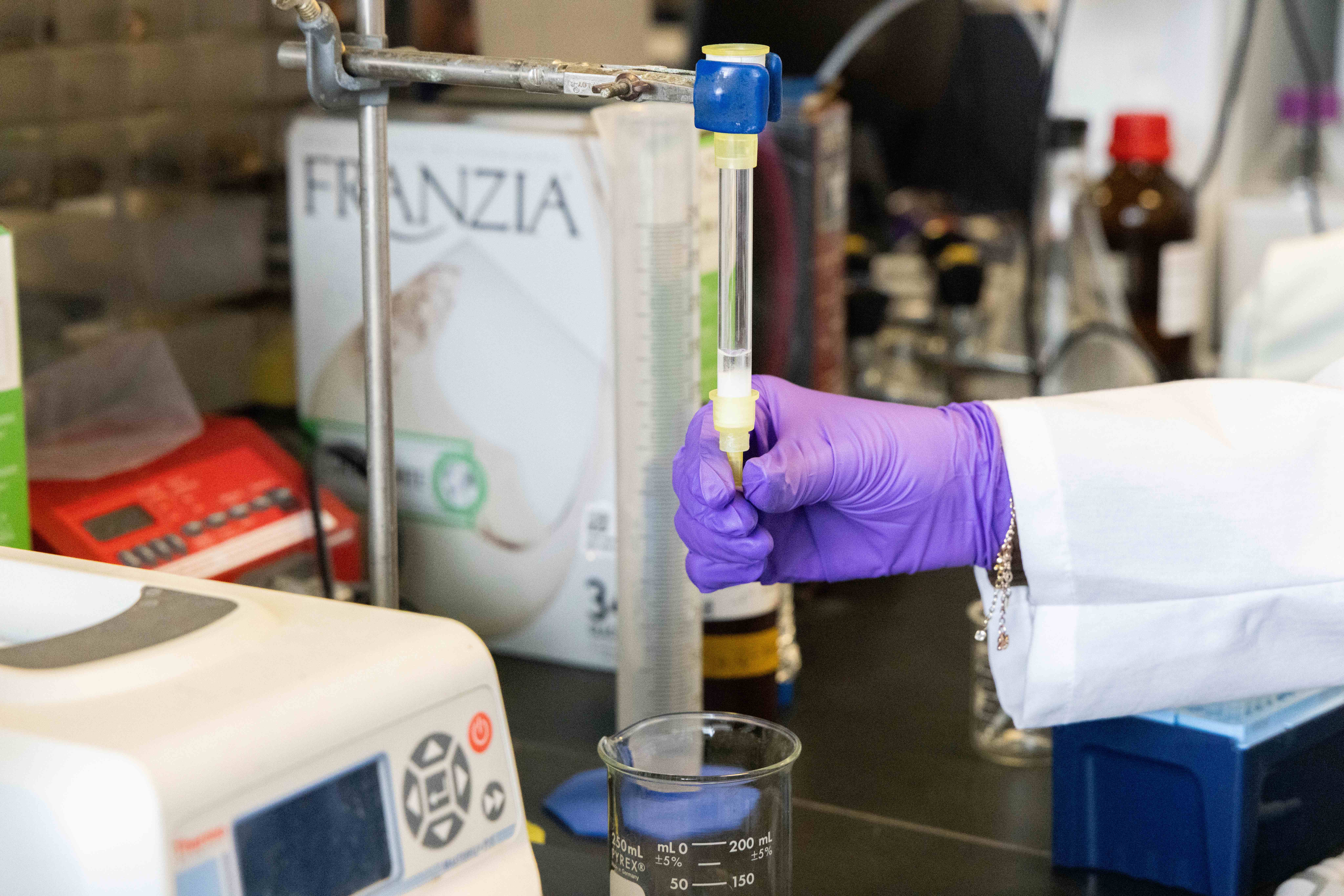 Purple-gloved hand using pipette to transfer clear liquid into beaker in lab; wine-box logo visible
