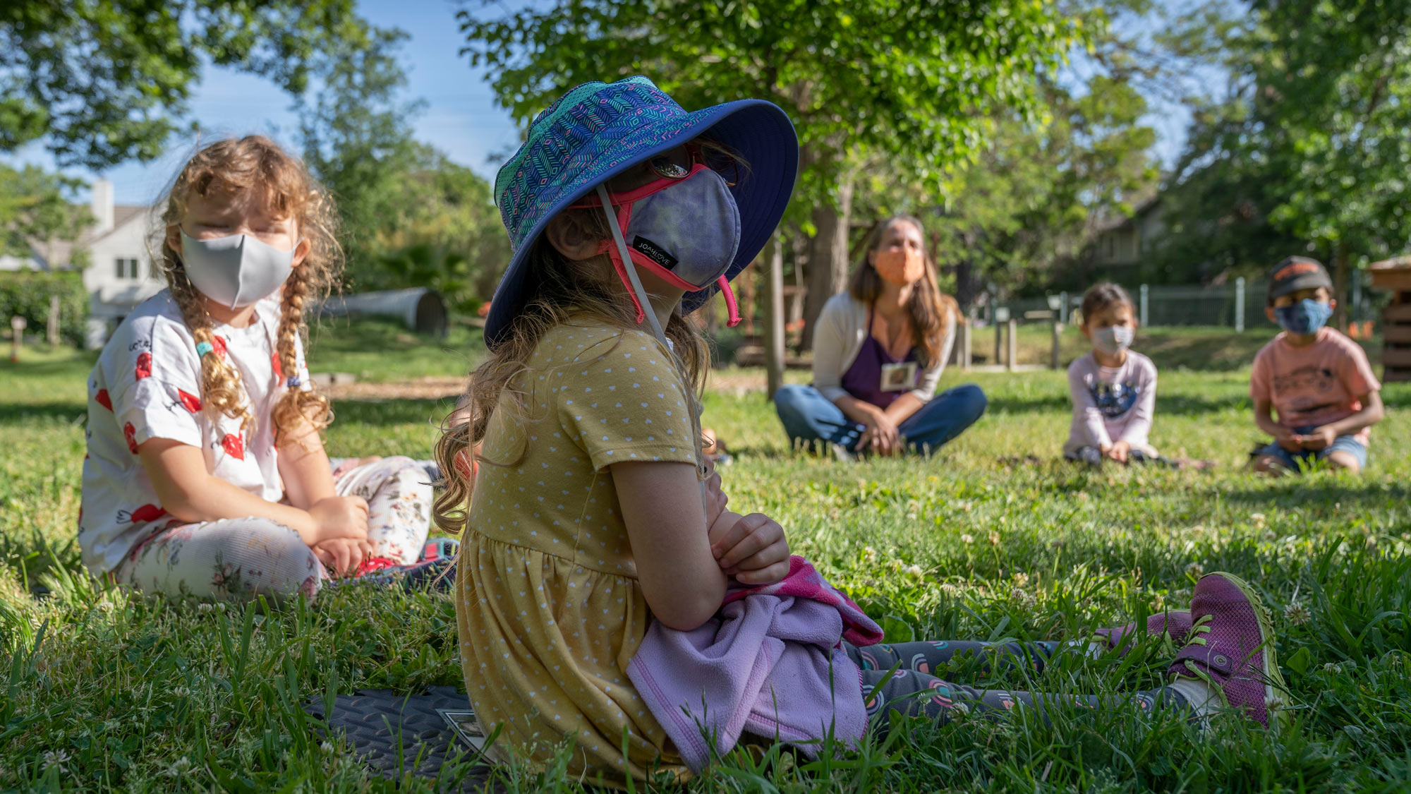 Toddlers, wearing face masks, sit in a socially-distanced circle on the grass.