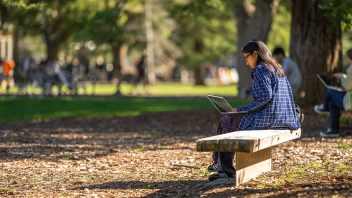 A student looks at her laptop while sitting on a bench outside.