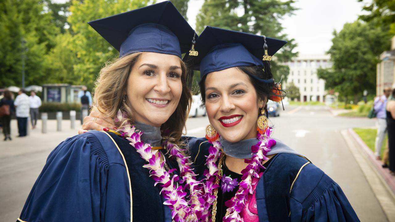 Two female graduates standing together in blue caps and gown.