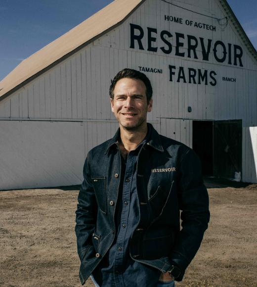 Young man in denim jacket smiling with hands in pockets in front of white Reservoir Farms barn