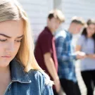 A teenage girl looking downward while standing apart of from a crowd of teens.