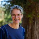Smiling person with short gray curly hair and glasses in blue sweater outdoors