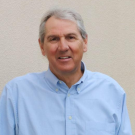 Smiling man in a light blue shirt standing against a neutral background.